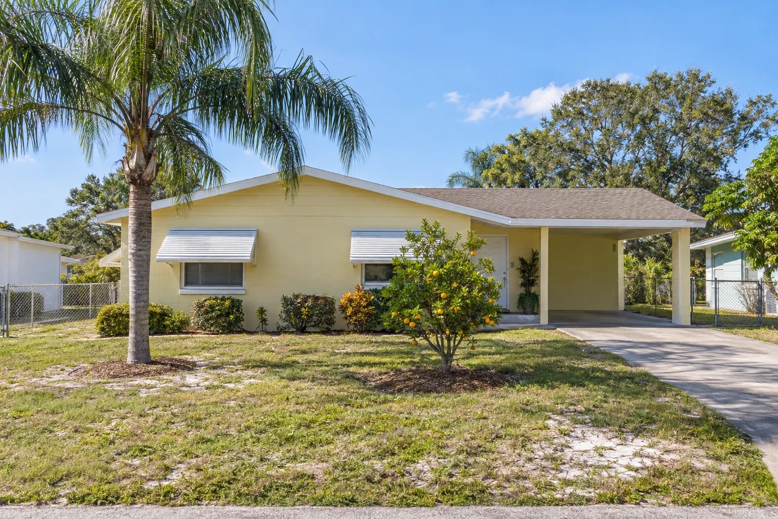 A one-story pale yellow block home in Pinellas Park with a low gable roof, white aluminum awnings over the windows, a single carport, and a palm in a sunny front yard