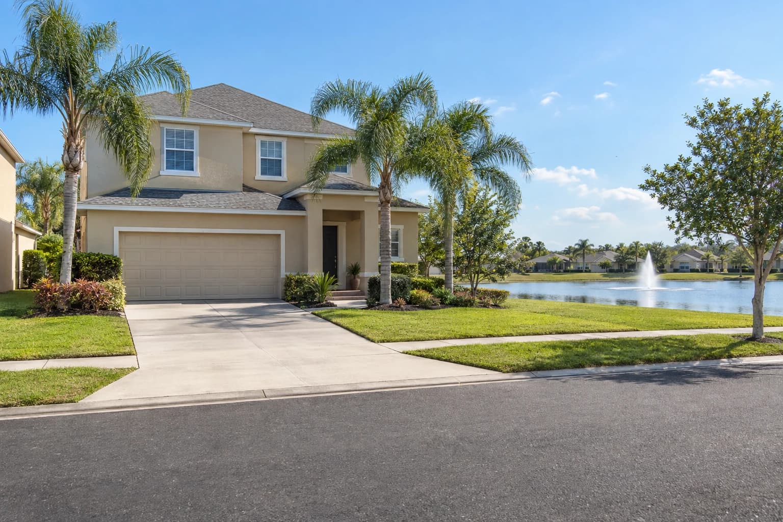 Two-story tan stucco home with a gray shingle roof, a two-car garage, and young palms beside a retention pond on a Riverview street