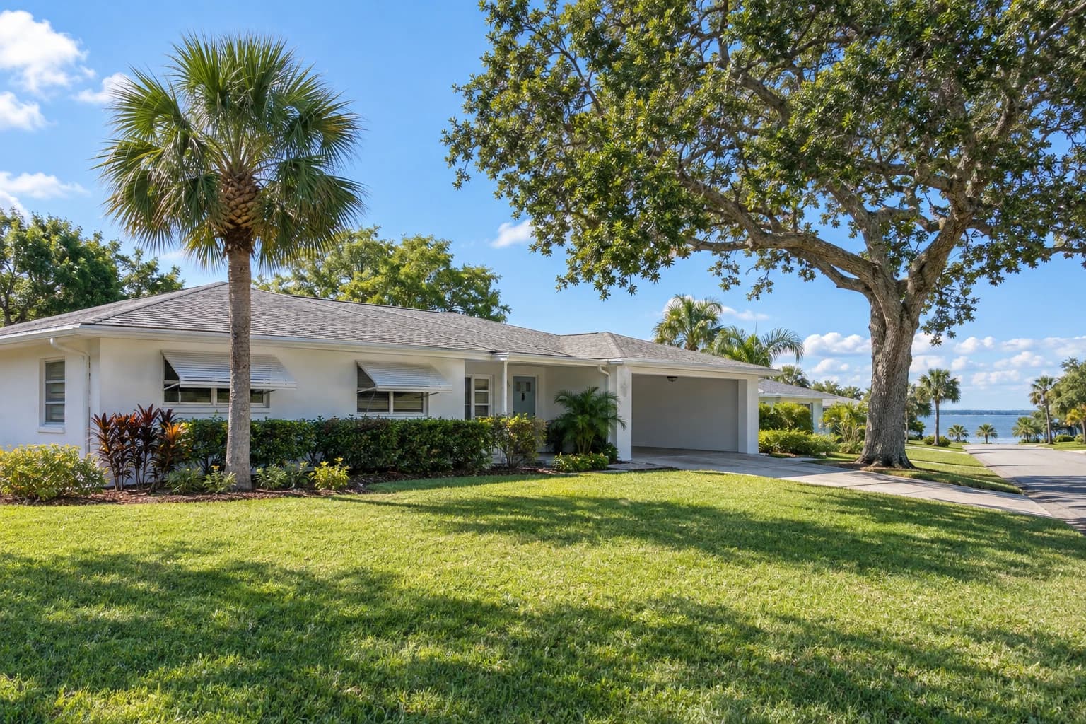 A one-story 1960s concrete block ranch home with white painted stucco, a low hip roof, a carport, and a cabbage palm and live oak in the front yard on a quiet Rockledge street