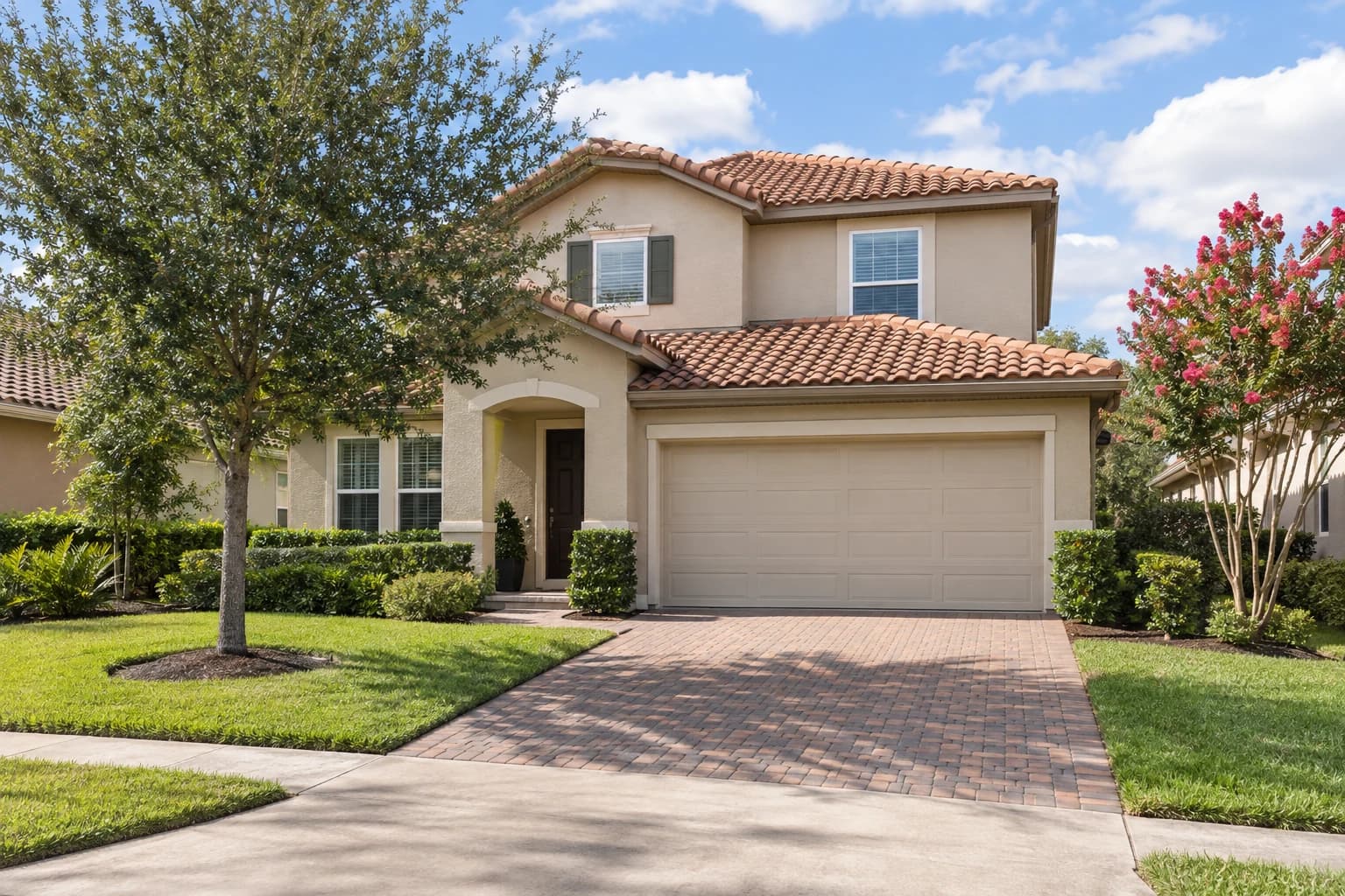A two-story stucco home from the 2000s with a barrel tile roof, a paver driveway, and a young oak in the front yard on a quiet Winter Garden street