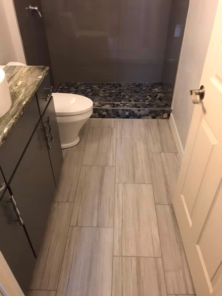 Remodeled bathroom with gray plank tile flooring, a curbed shower with a pebble stone floor, and a dark vanity with a granite top