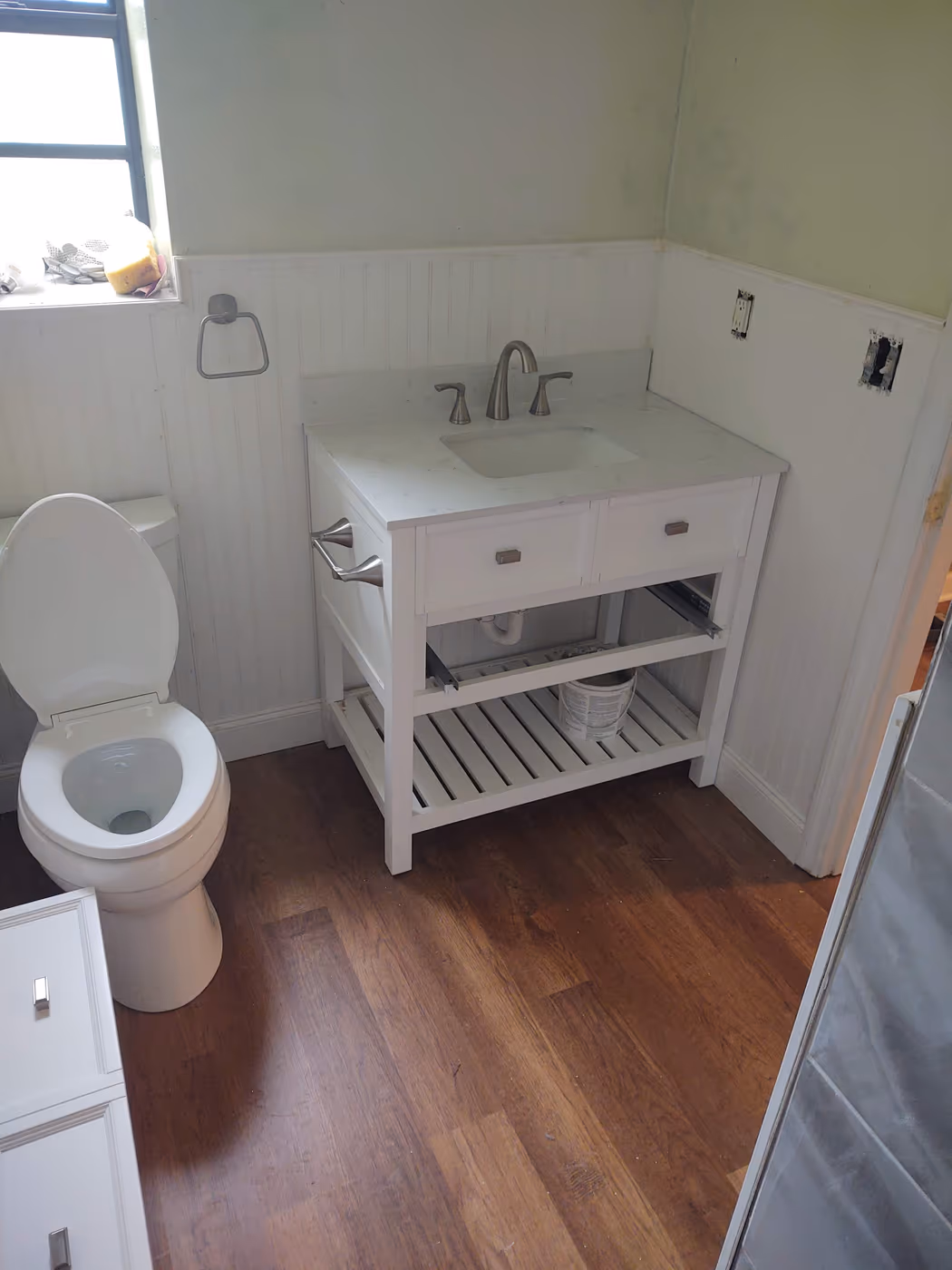 Remodeled bathroom by our team with a white toilet beside a new white vanity, beadboard walls, and wood flooring