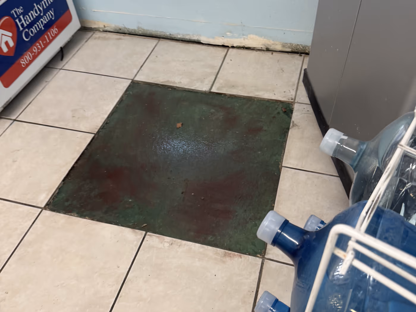 Office break room with a failed tile patch over bare concrete and crumbling drywall at the base of the wall